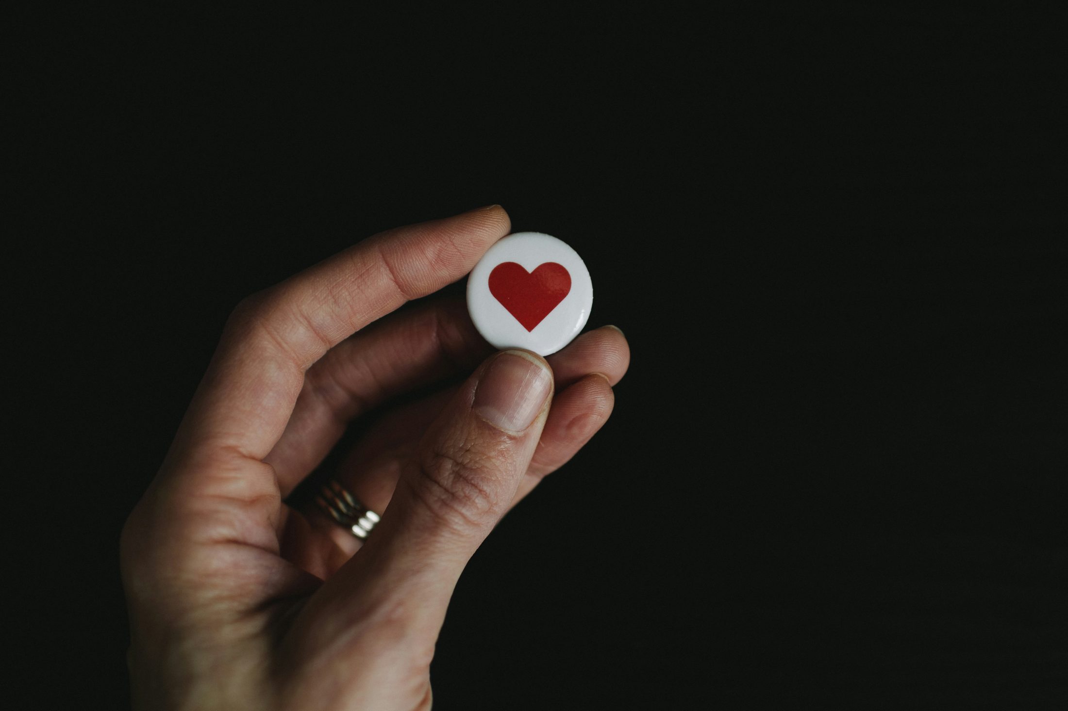 A hand holding a small white pin badge with a red heart, photographed against a black background.